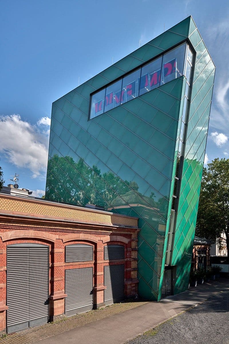 Ein grüner Turm ist zu sehen. Der Turm ist schief. Ganz oben ist ein langes Fenster. Das ist die Kunsthalle von außen.